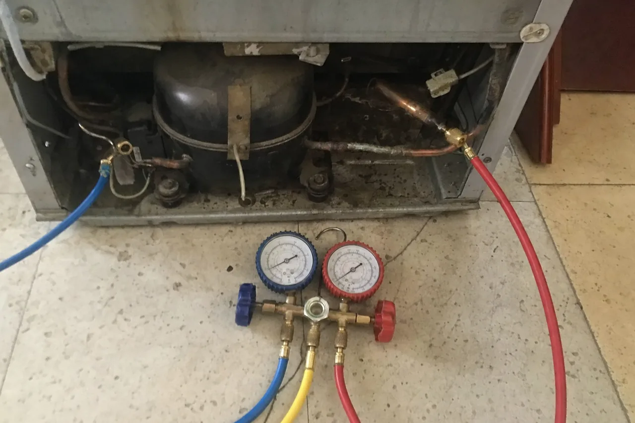 A manifold gauge set standing on the floor with its hoses connected into the compressor compartment of a refrigerator