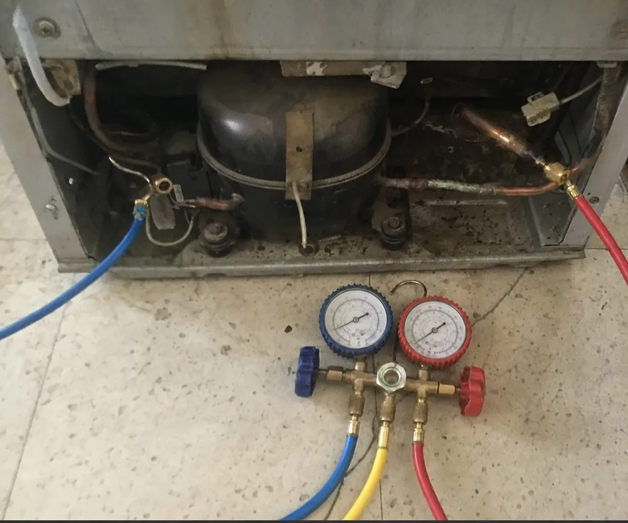 Gauge hoses running from a manifold set into the machine compartment at the base of a refrigerator